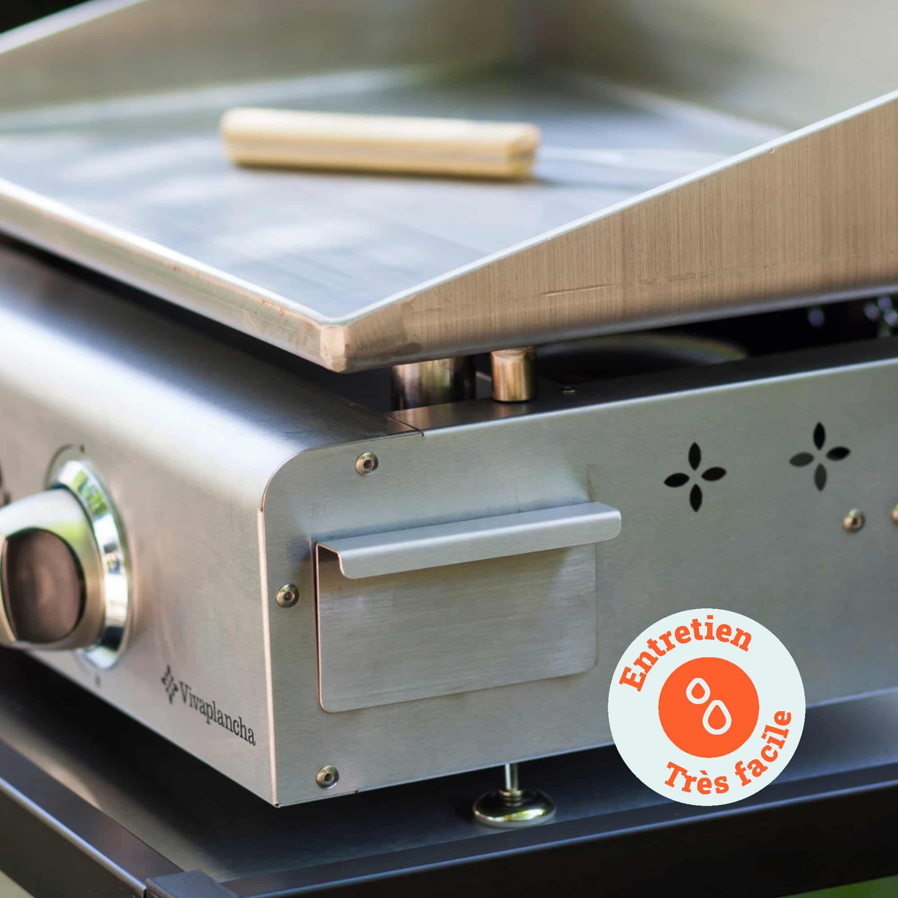 Primer plano de la plancha Vivaplancha mostrando el recogedor de grasa de acero inoxidable y una herramienta de limpieza colocada sobre la placa, con una insignia que dice “Mantenimiento muy fácil”.