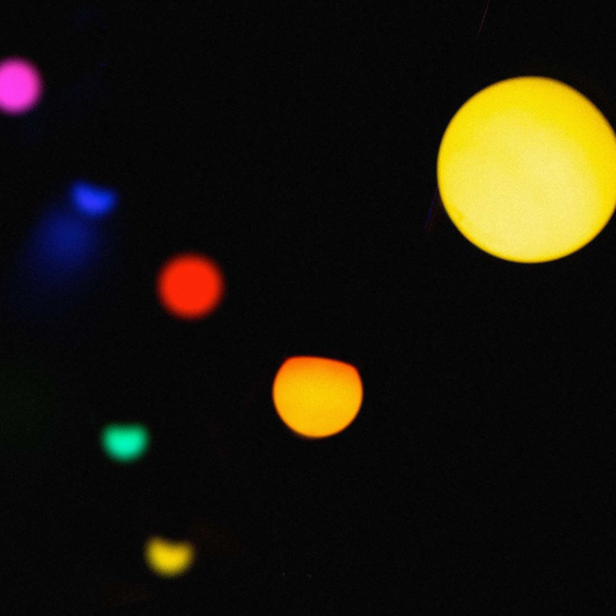 Guirnalda de luces exterior multicolor desenfocada en la noche, con puntos de luz amarillos, naranjas, rojos, verdes, azules y violetas sobre un fondo oscuro.