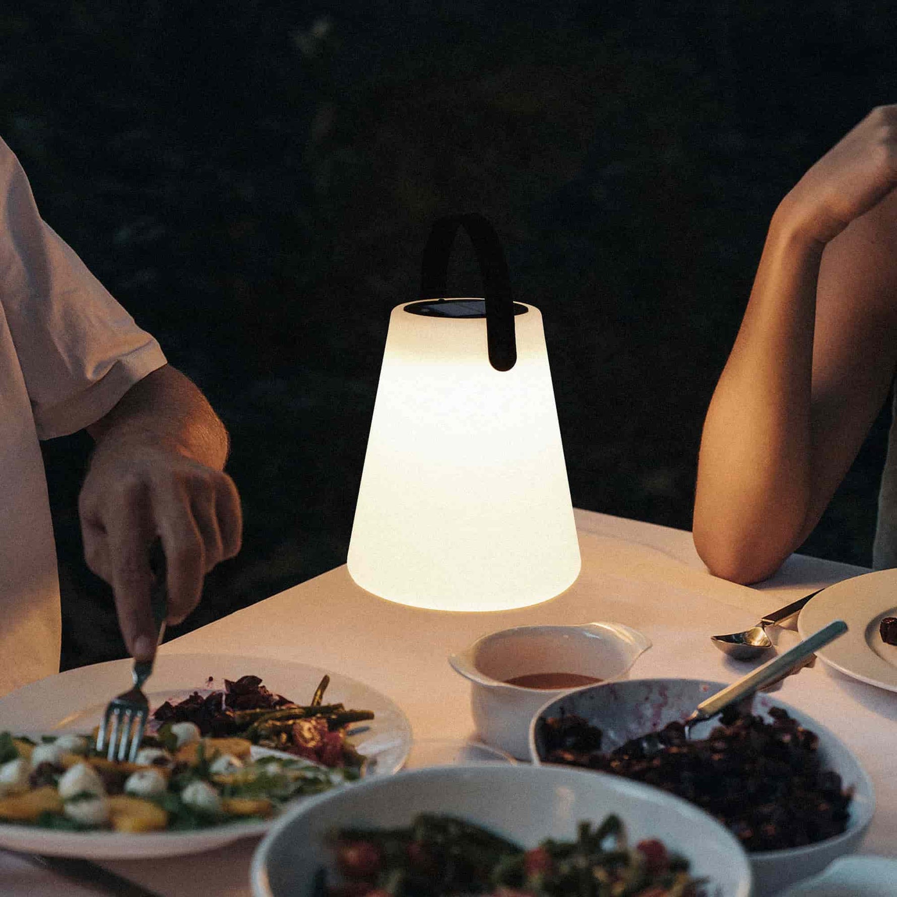 Lampe conique solaire allumée, diffusant une lumière chaude sur une table dressée pour un dîner en extérieur, entourée de plats et de convives.