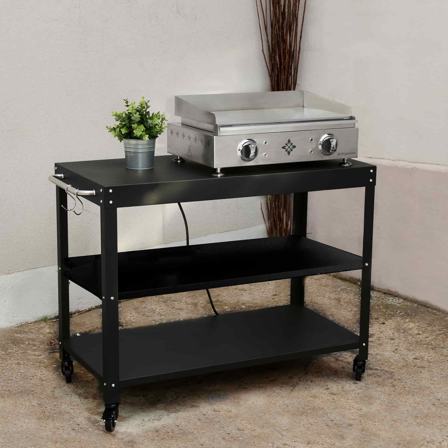 Carro negro para plancha con tres niveles de almacenamiento, presentado en exterior con una plancha eléctrica de acero inoxidable colocada en la estantería superior y una pequeña maceta con planta decorativa.