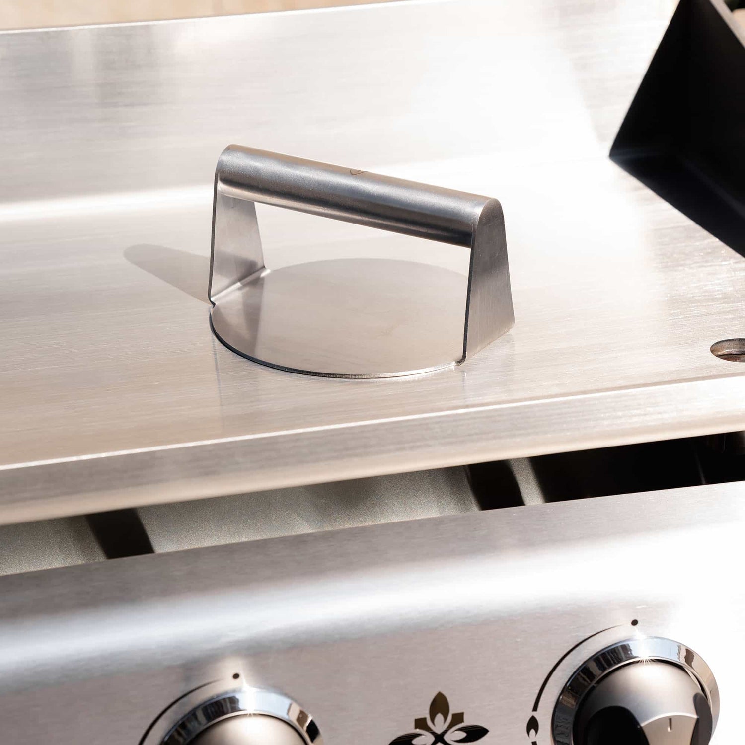 Presse à smash burger en inox posée sur la plaque d’une plancha inox à trois brûleurs, présentée en gros plan en extérieur.